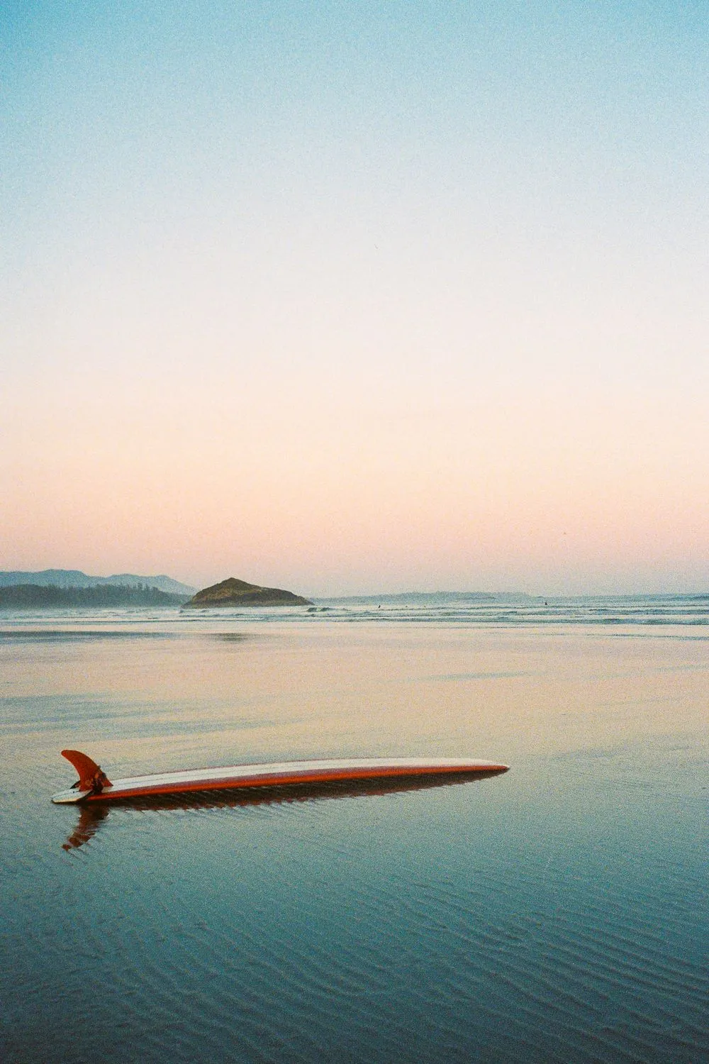 Surfboard lying in shallow water at dusk, with a small island on the horizon