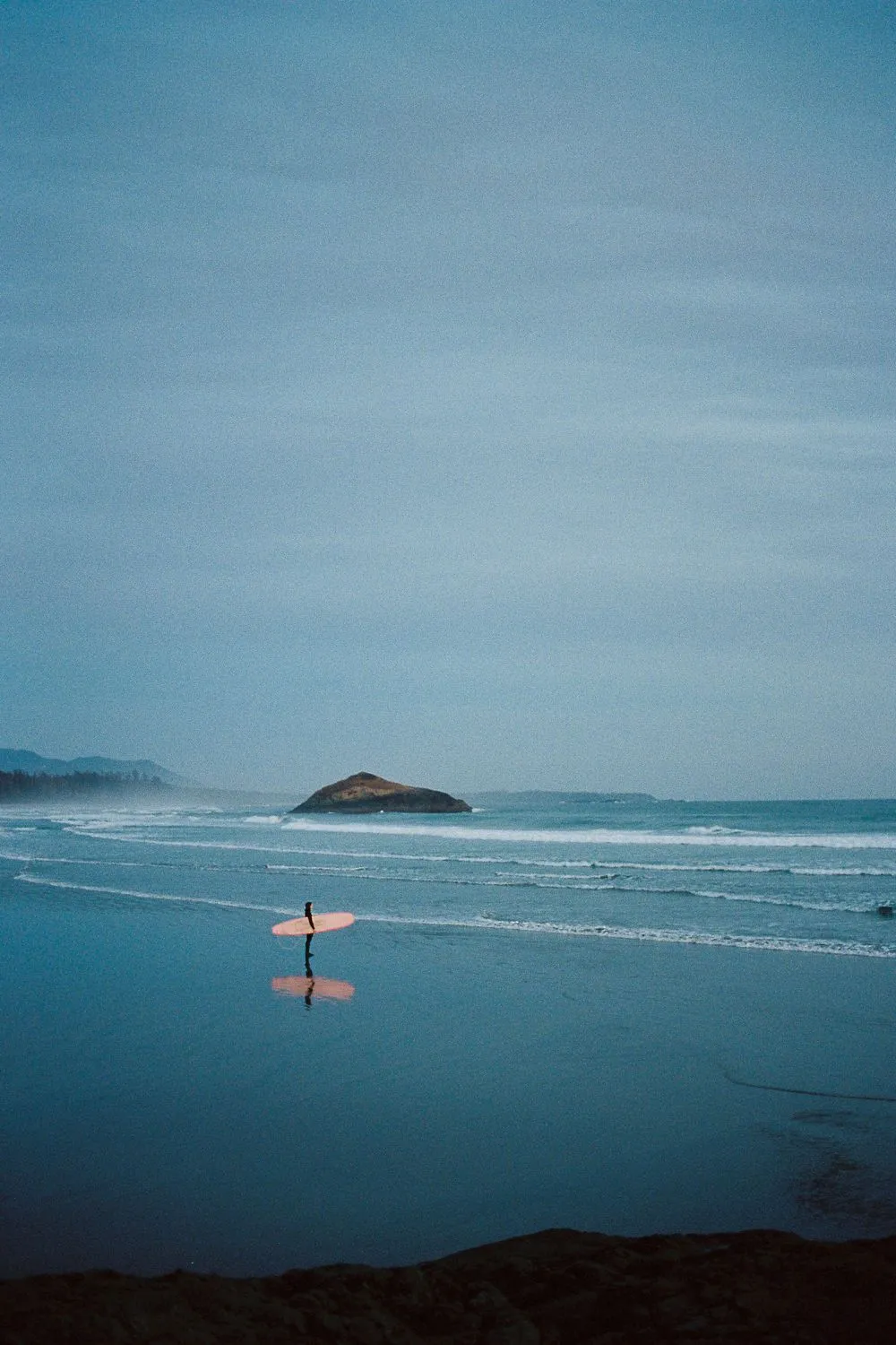 Surfer carrying a board along a calm beach towards a small island