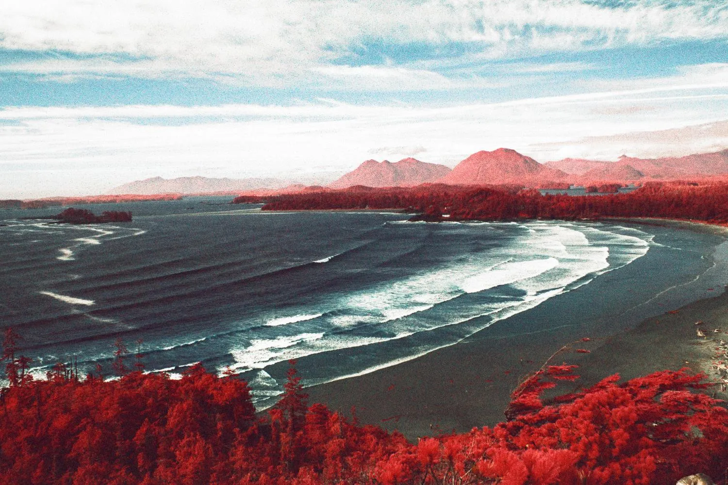 Infrared view over a curving surf beach, red forest in the foreground and red-tinted mountains beyond